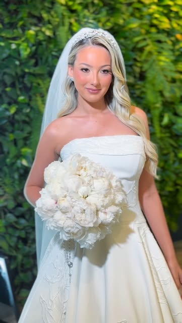 Noiva com véu, cabelo loiro em ondas e buquê de peônias brancas